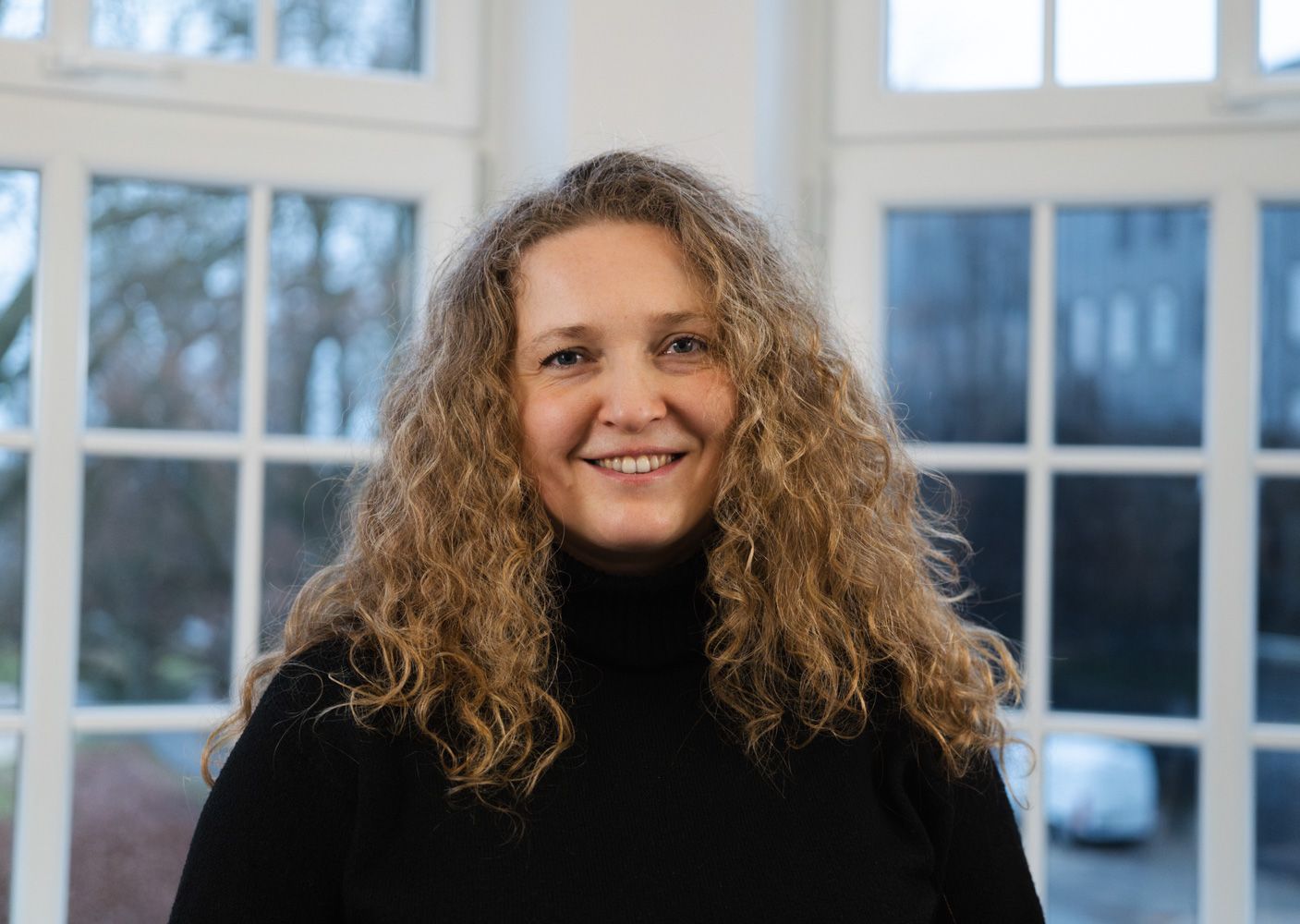 IDALABS-Mitarbeiterin Patrycja steht vor einer Fensterfront und lacht in die Kamera. Sie hat dunkelblonde Locken und trägt einen schwarzen Rollkragenpullover.