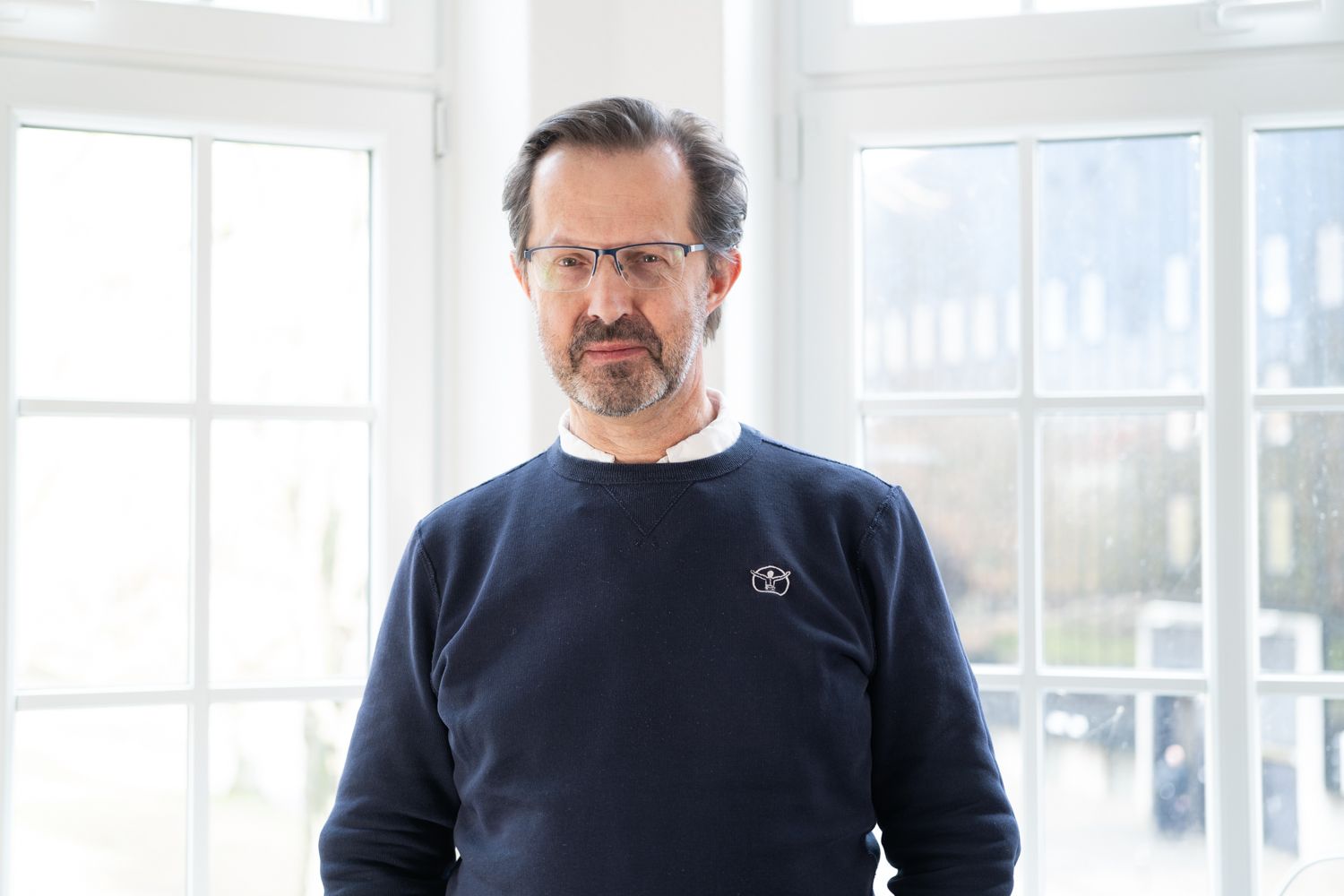 Ein Mann mit braunen Haaren und Dreitagebart lächelt in die Kamera. Er trägt Brille und ein weißes Hemd.