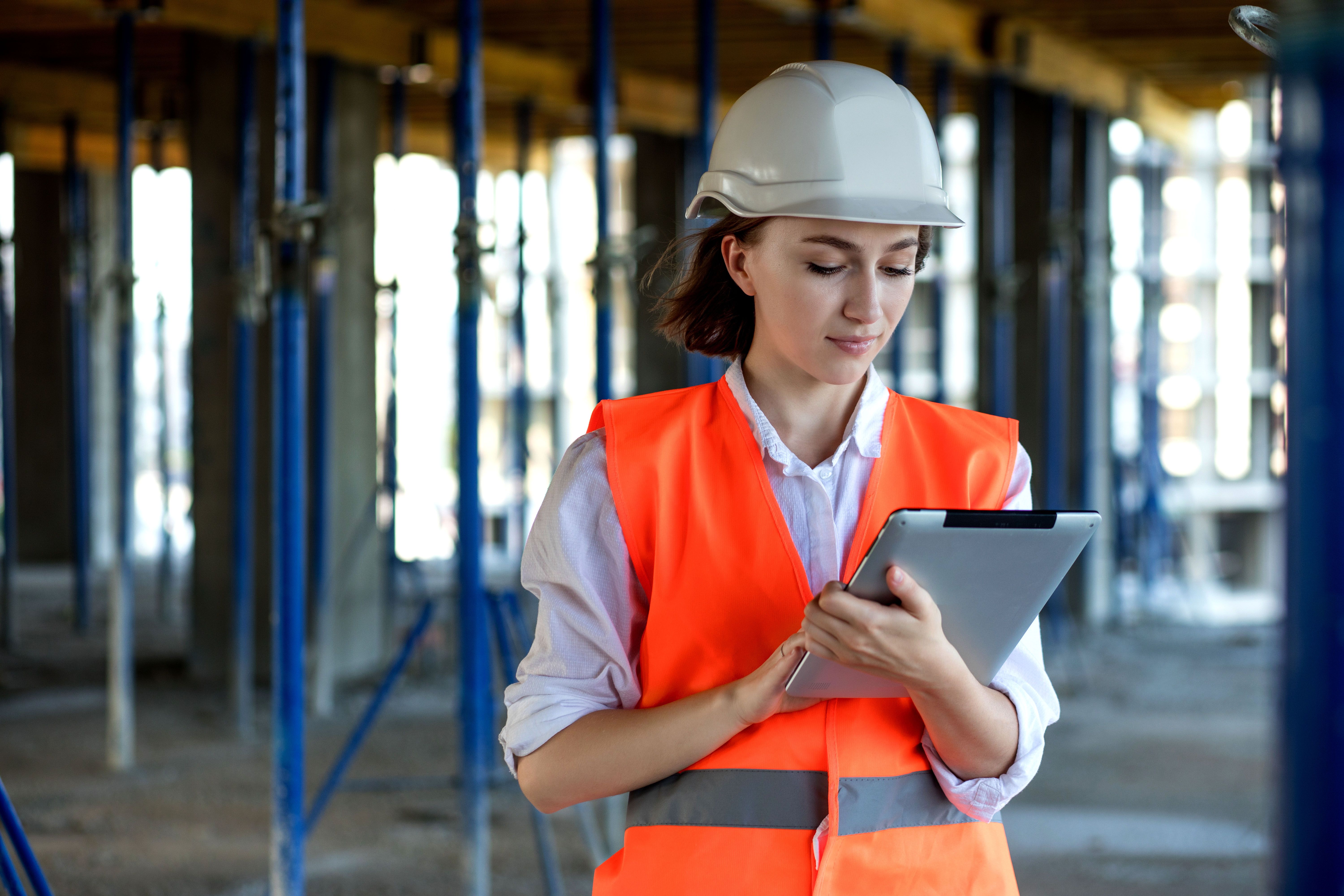  Eine junge Frau, die eine orangefarbene Warnweste und einen weißen Sicherheitshelm trägt, steht vor einer halbfertigen Baukonstruktion. Sie arbeitet an einem Tablet.