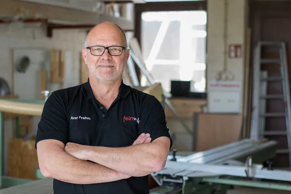 Arne Frahm is standing in a workshop. He is wearing a dark T-shirt with the company logo and a pair of glasses.