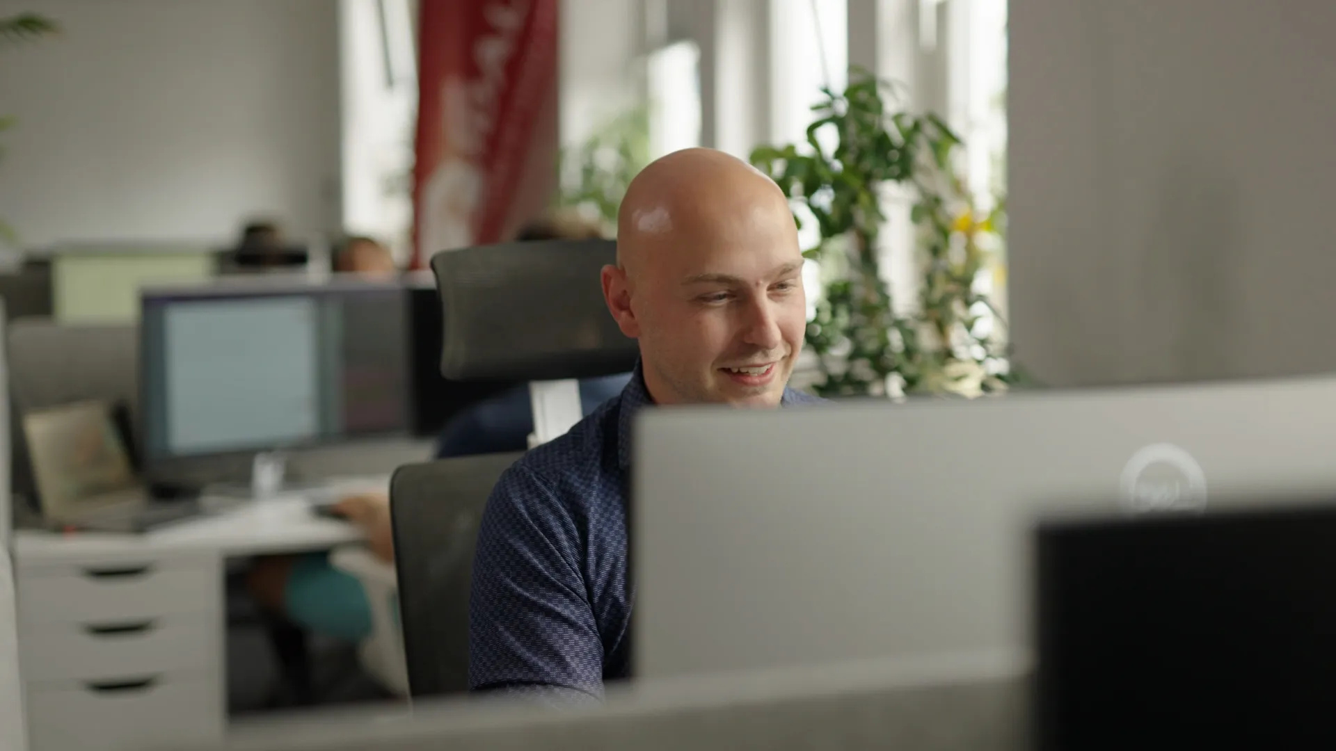 Alexander Westen, Geschäftsführer bei IDALABS, sitzt an einem Schreibtisch und arbeitet am Computer. Im Hintergrund ist das IDA-Büro mit weiteren Teammitgliedern an ihren Arbeitsplätzen zu sehen.