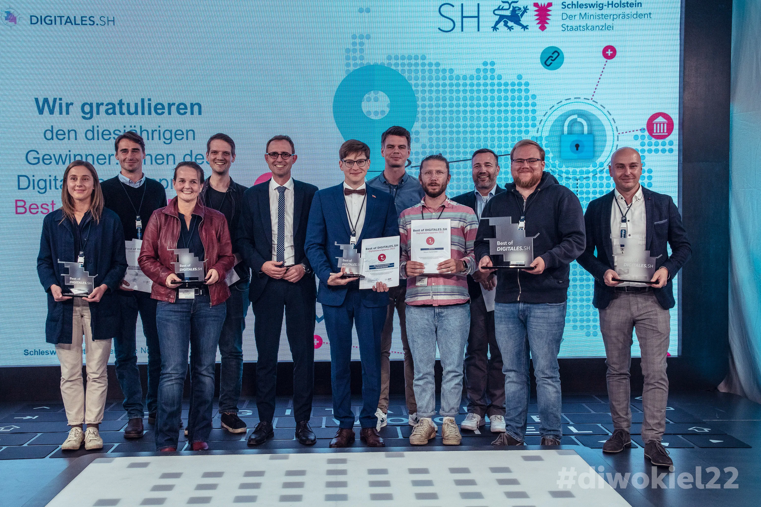 Vor einer beleuchteten Leinwand stehen 11 Männer und Frauen, darunter die IDALABS-Gründer Alexander Westen und Tobias Gürtler. Sie zeigen stolz ihre Trophäen für die Auszeichnung zur “Best of Digitales S.H.”.