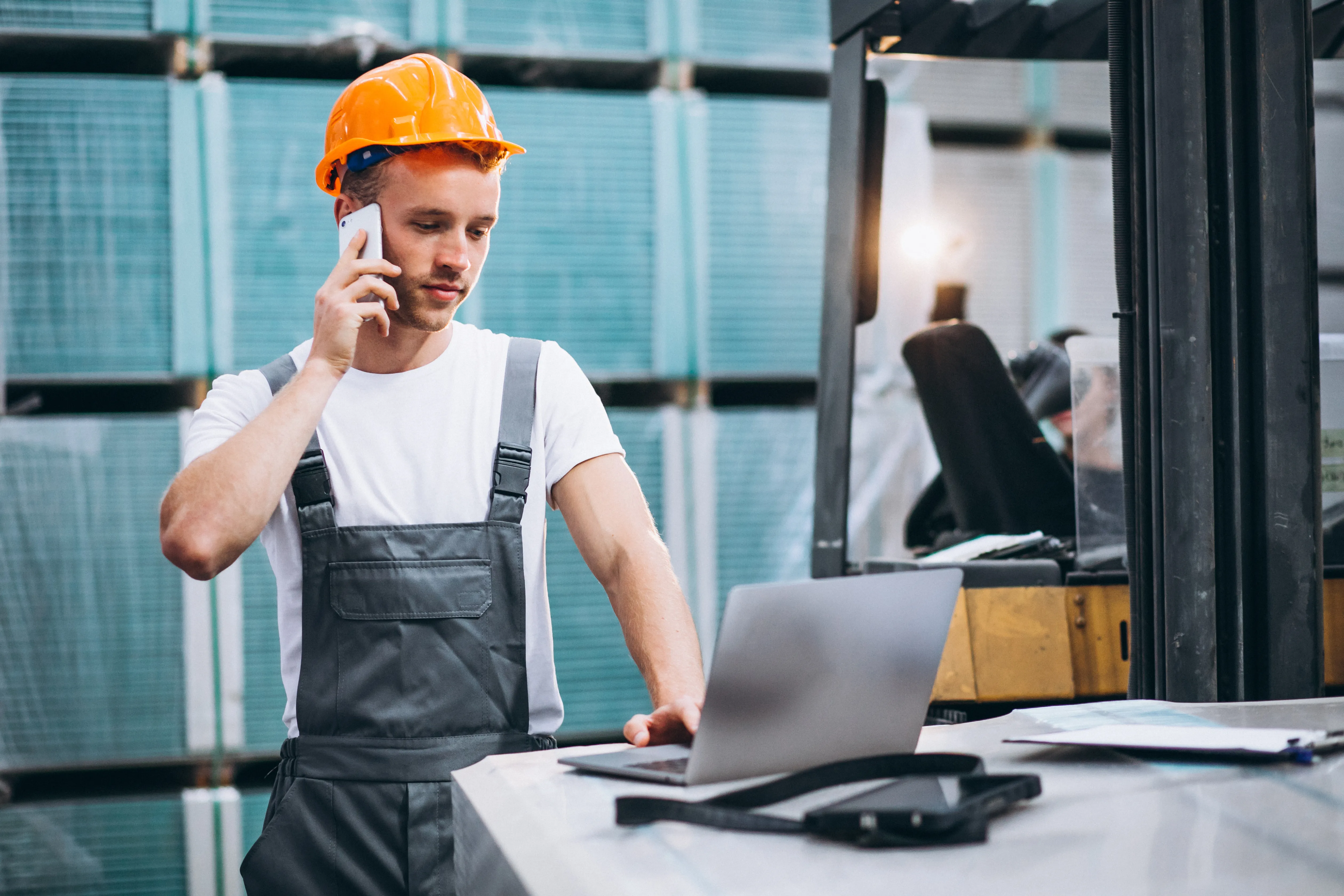 In einem Warenhaus steht ein Mitarbeiter an einem Tisch vor seinem Laptop. Er trägt einen orangefarbenen Sicherheitshelm und hält sich zum Telefonieren ein Smartphone ans Ohr. Neben ihm ist ein Gabelstapler zu sehen.