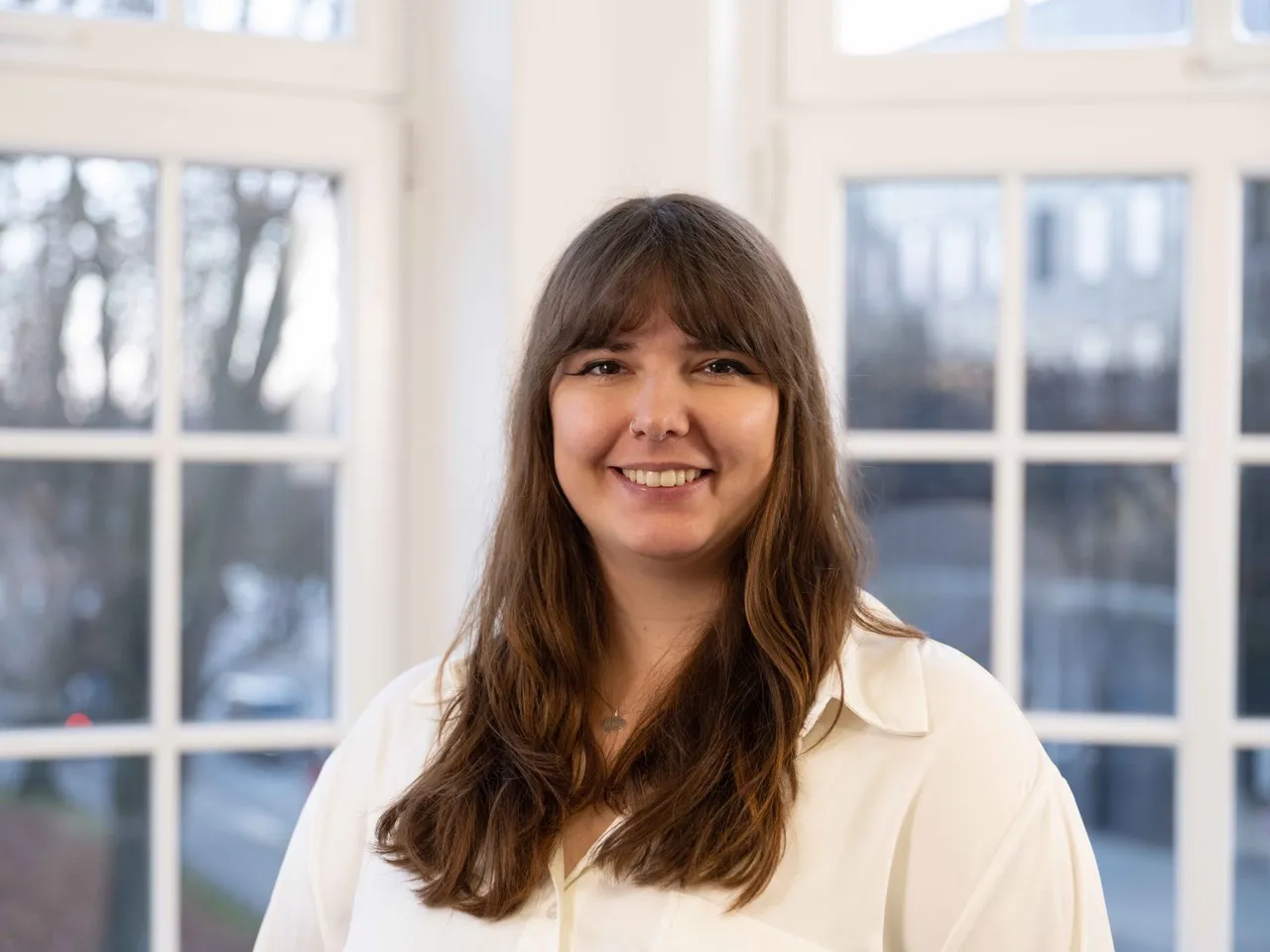 Svenja Cichon steht vor einer Fensterfront in einem Büro. Sie trägt ihre Haare lang mit Pony und hat ein weißes Hemd an