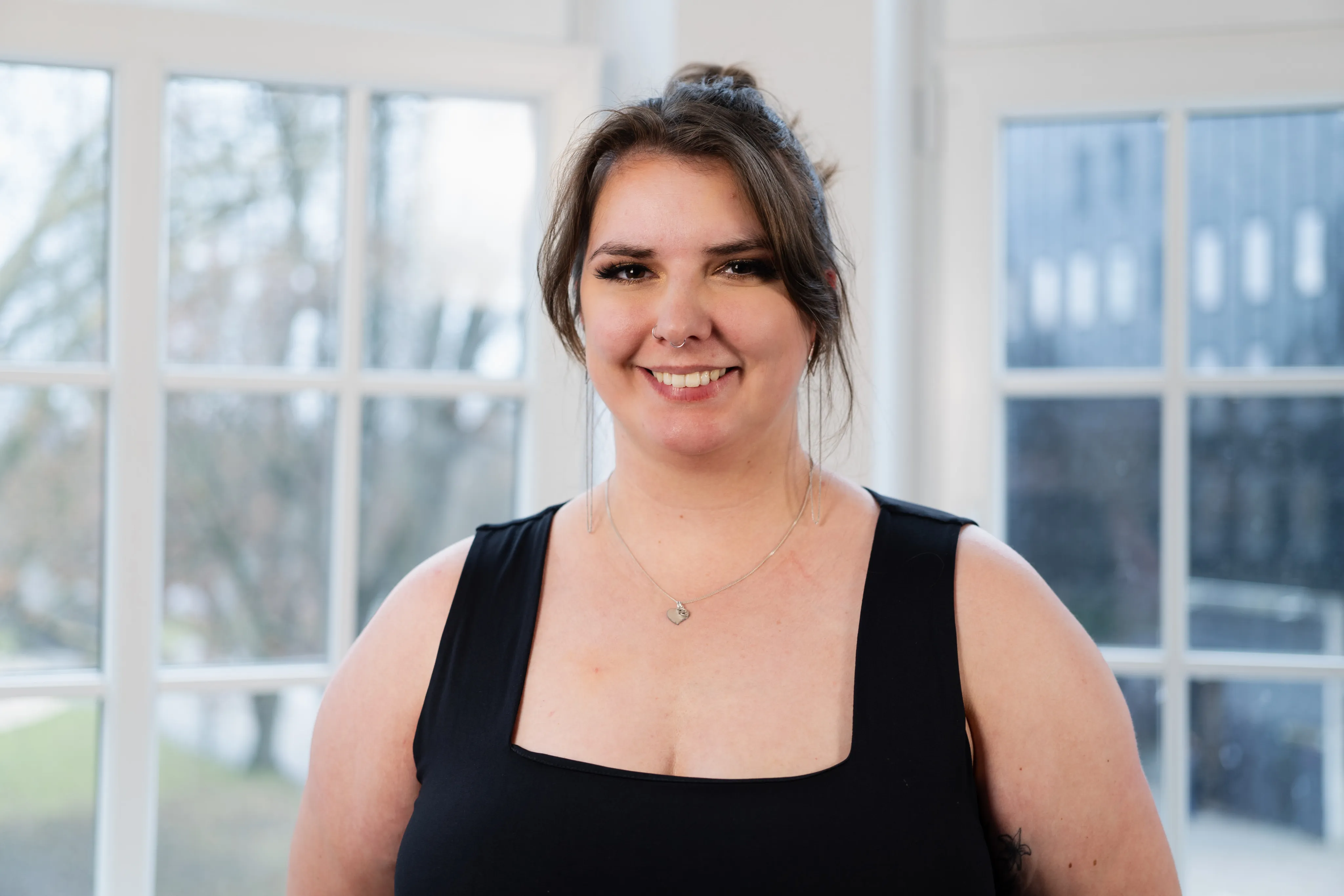 Svenja Cichon steht vor einer Fensterfront in einem Büro. Sie trägt ihre Haare im Dutt mit Pony und hat ein schwarzes Oberteil an.