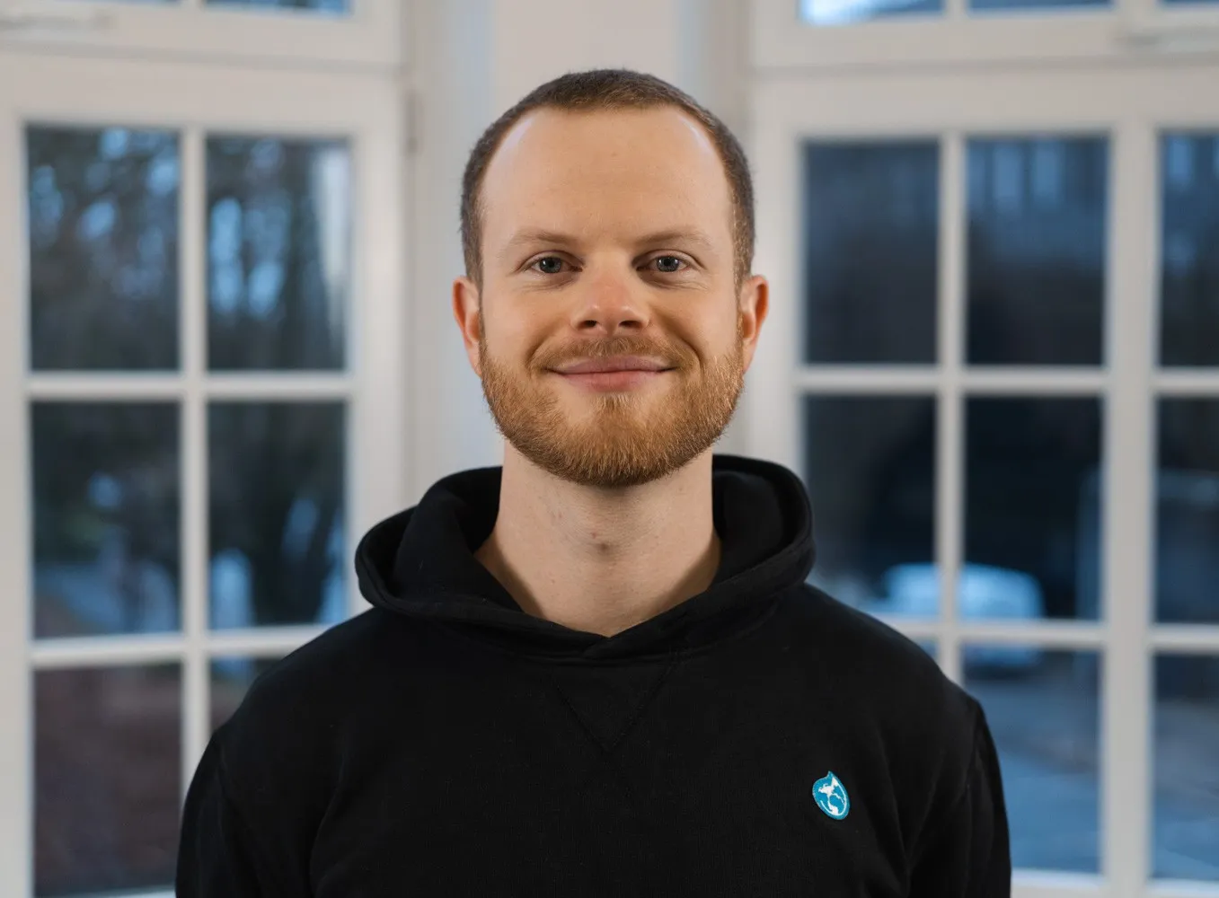 Joshua Winkelmann schaut freundlich in die Kamera. Er trägt einen schwarzen Kapuzenpullover und seine rötlich-braunen Haare kurz geschnitten
