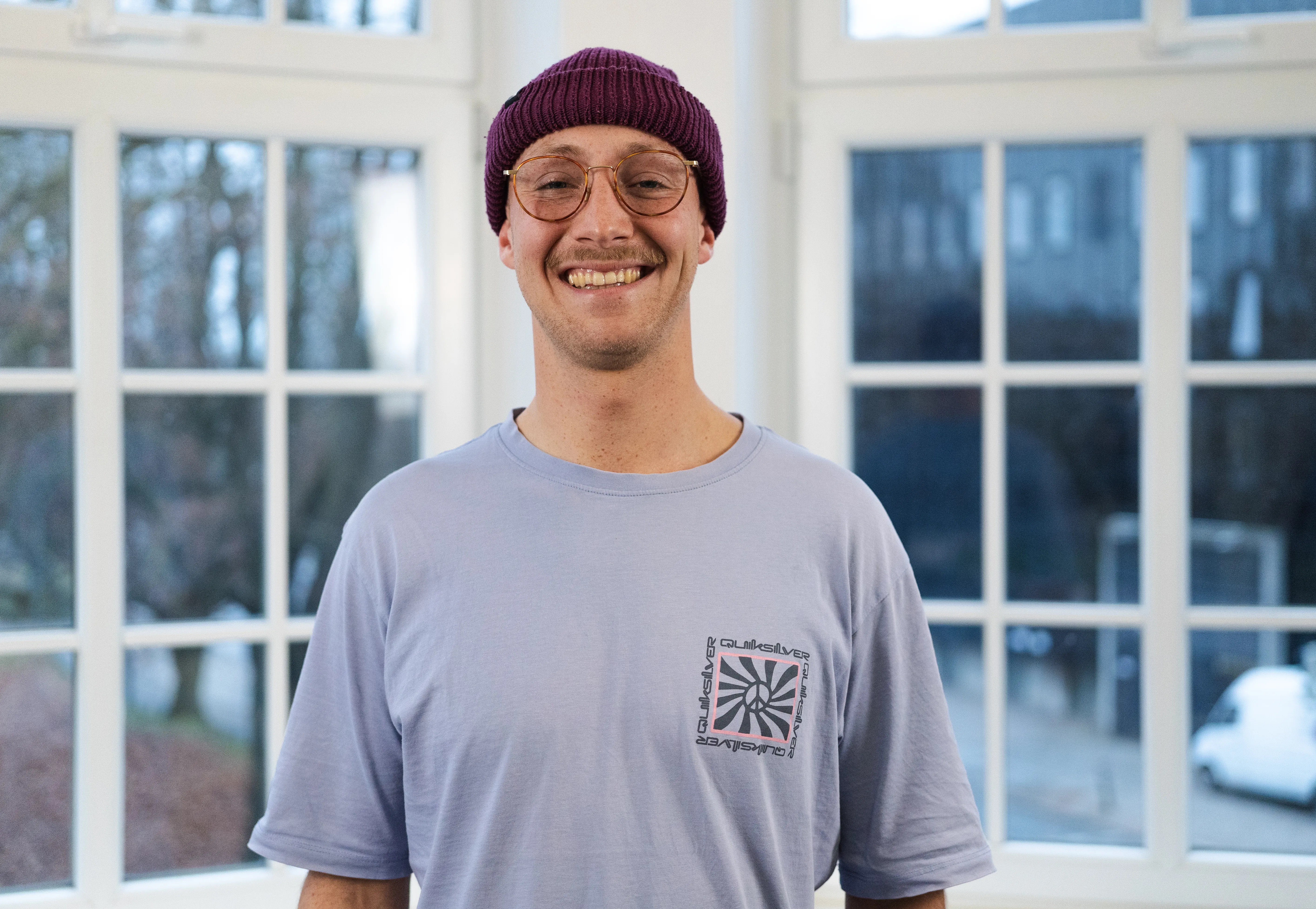 IDALABS-Mitarbeiter Mirco steht vor einer Fensterfront und schaut aufmerksam in die Kamera. Er trägt eine weinfarbene Mütze, Brille, einen Schnurrbart und ein hellblaues T-Shirt.