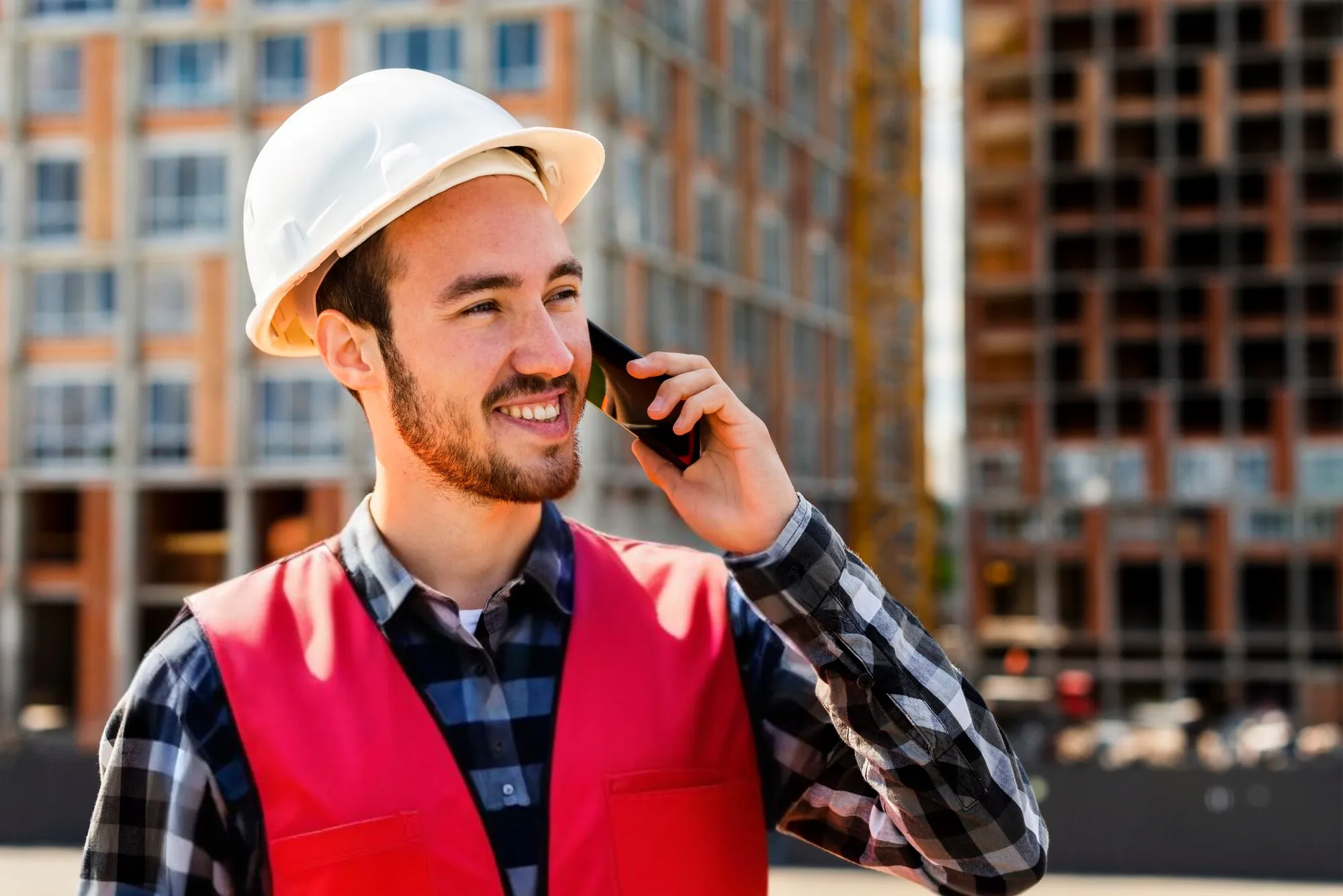 Haben wir Dein Interesse geweckt? Vor einem mehrstöckigen Gebäude mit Glasfront ist ein Mann zu sehen, der lächelnd ein Smartphone an sein Ohr hält. Er trägt ein kariertes Hemd sowie eine rote Warnweste und einen weißen Sicherheitshelm