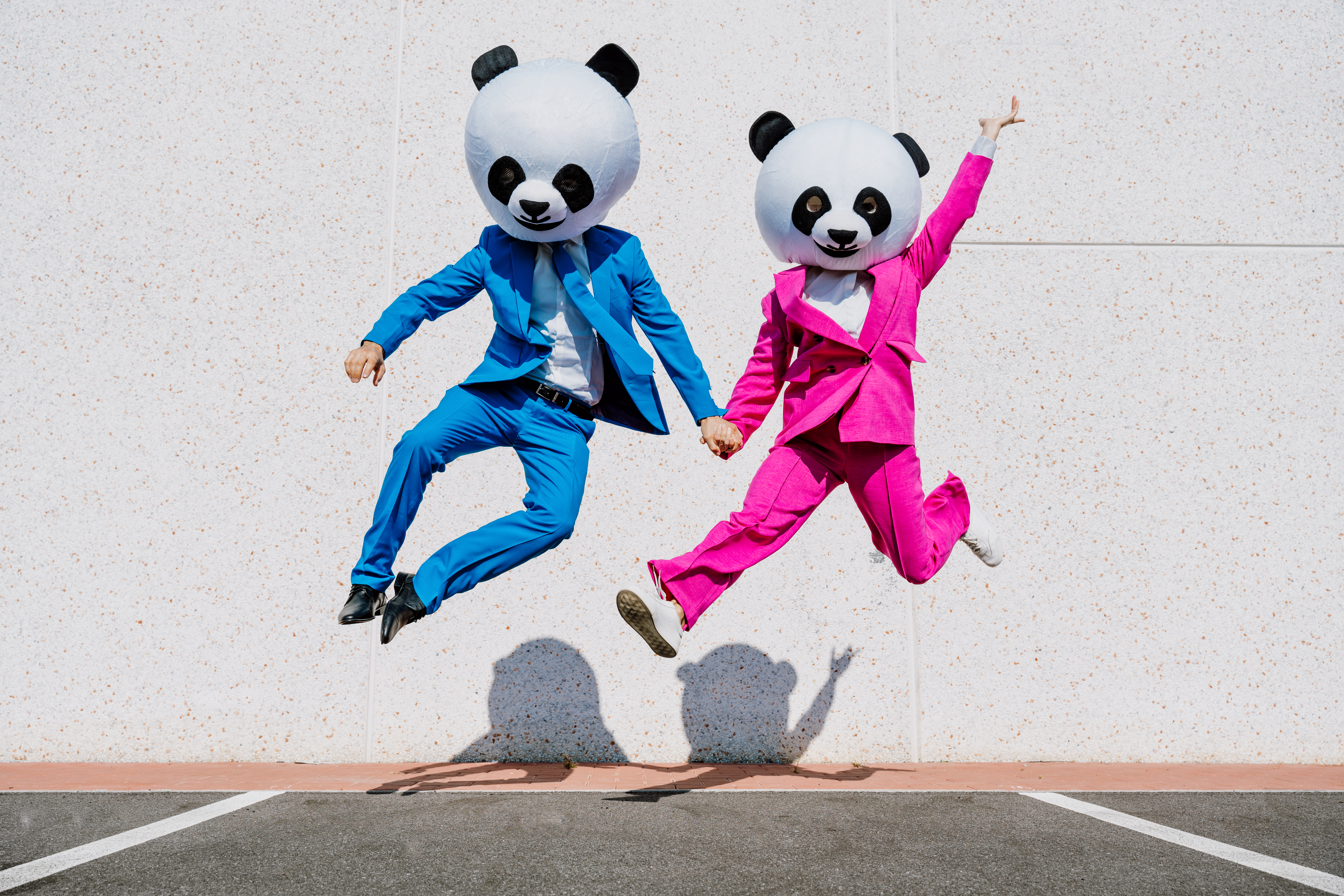 Zwei Menschen springen dynamisch in die Höhe und halten sich dabei an den Händen. Der eine trägt einen blauen Anzug mit blauer Krawatte, der andere einen pinken Anzug. Beide tragen übergroße Pandamasken.